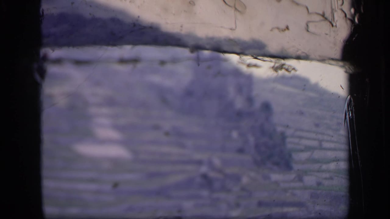 1949 8mm home-movie frame from Ireland showing terraced hillside fields and a distant castle-like ruin through dark window bars.