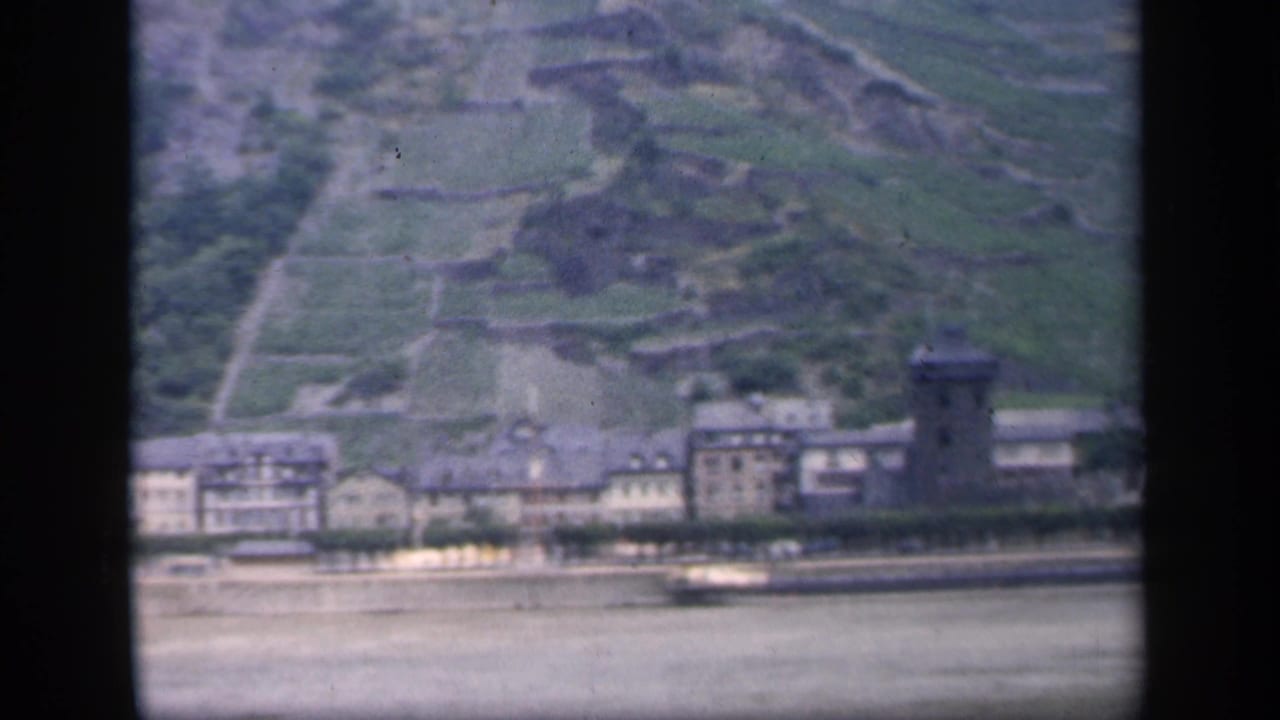 Soft 8mm view of riverside buildings, a tower, and terraced green hillside across the water.