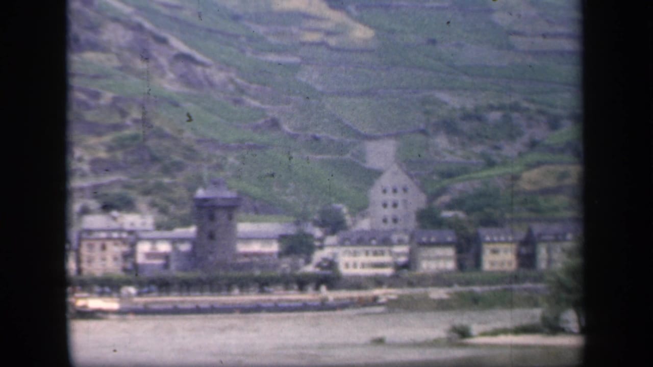 Mid-century 8mm frame of a waterfront settlement with a tower, pale road, and cultivated hillside, with Ireland noted in clip metadata.
