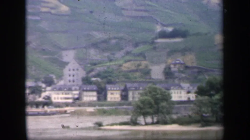 1949 home-movie frame showing trees by the river, pale waterfront buildings, and a steep terraced hillside above.