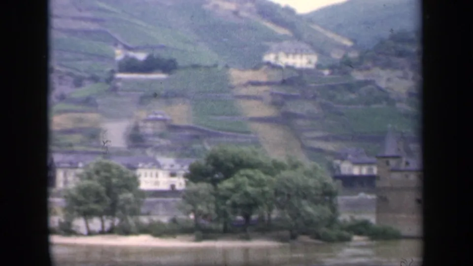 Soft 8mm frame from the end of the pan, showing riverbank trees, distant buildings, hillside terraces, and a partial tower.