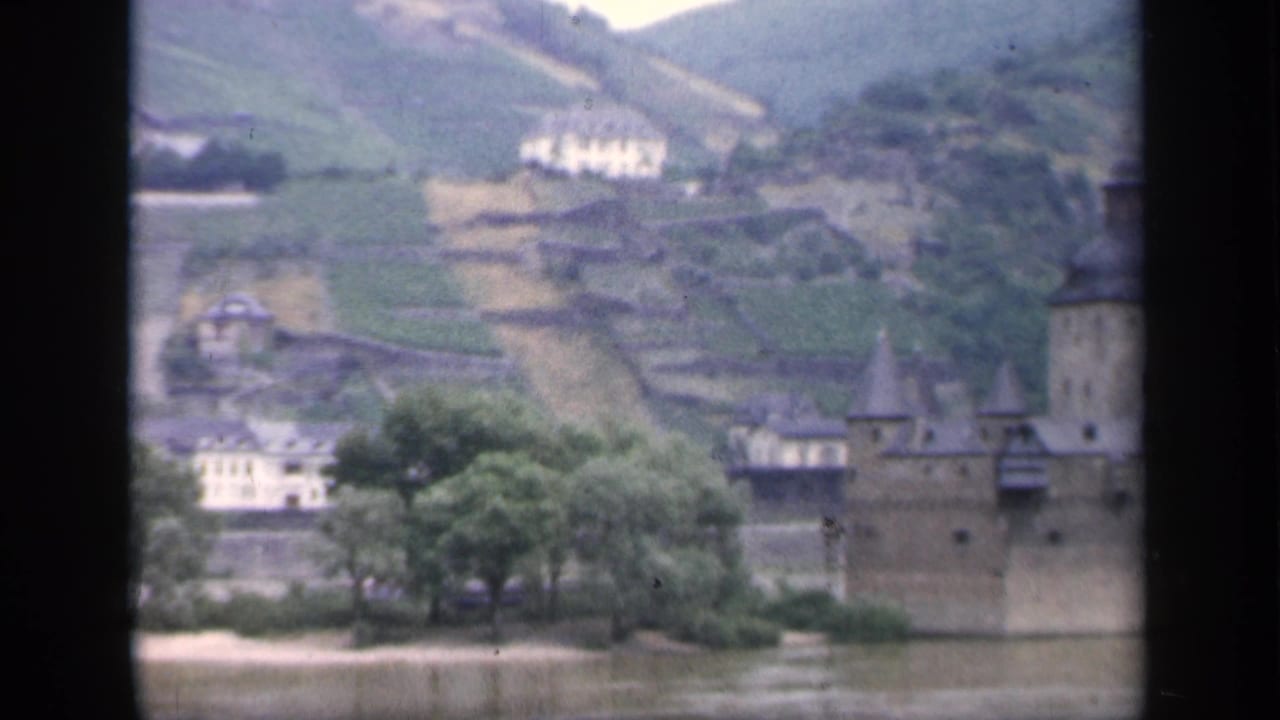 1949 8mm view of a riverside castle in Ireland, with stone walls, a tower, trees, terraced hills, and distant buildings.