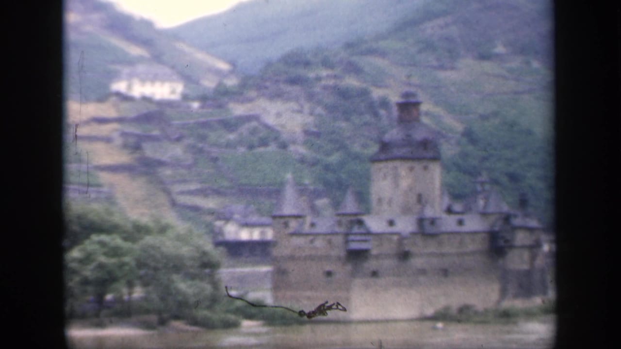 Soft 8mm frame of an Irish riverside castle, showing the central tower, turrets, riverbank trees, and terraced hillside beyond.