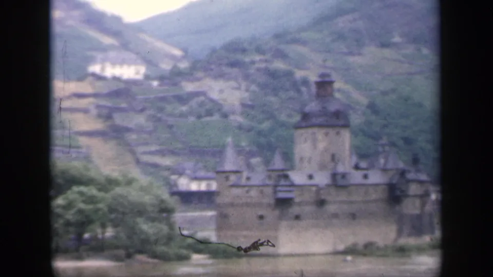Soft 8mm frame of an Irish riverside castle, showing the central tower, turrets, riverbank trees, and terraced hillside beyond.