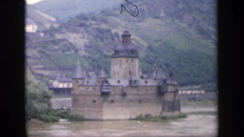 1949 home-movie frame showing the riverside castle in Ireland with a tall tower, smaller turrets, stone walls, trees, and hillside fields.