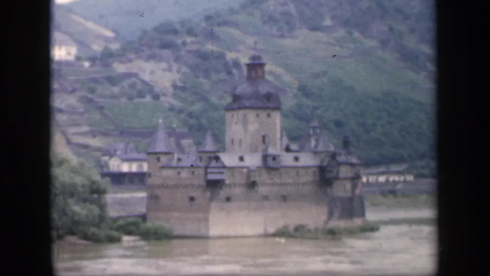 Vintage 8mm view of an Irish castle by a muddy river, with soft focus, visible film wear, hillside terraces, and pale buildings behind.