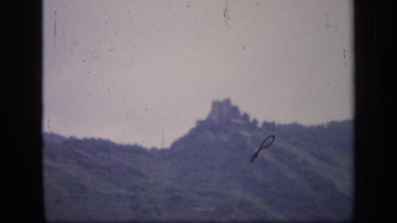 1949 Ireland 8mm frame showing a distant hilltop ruin above a tree-covered slope under a pale overcast sky.