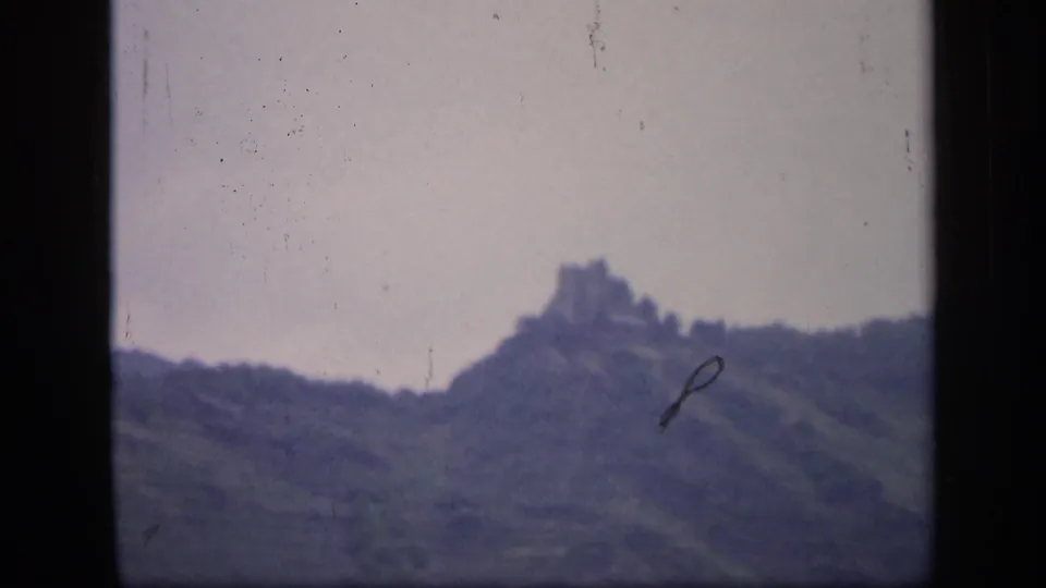1949 Ireland 8mm frame showing a distant hilltop ruin above a tree-covered slope under a pale overcast sky.