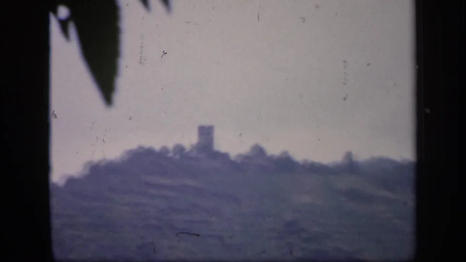 8mm frame from Ireland showing a distant hilltop tower or ruin, with a dark foreground leaf intruding from above.