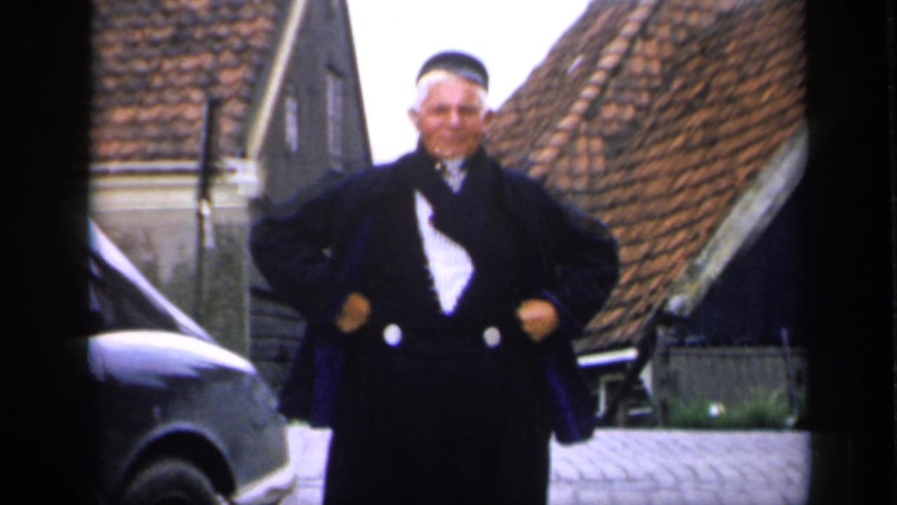 Man in dark coat and cap posing on a cobblestone street beside a rounded car.