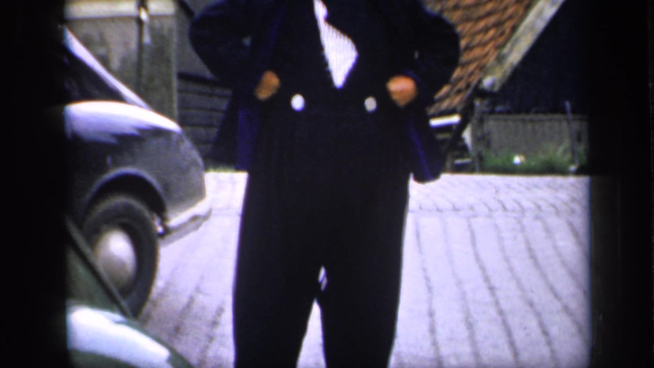 A cropped 1949 Ireland frame shows the man's dark coat and trousers beside a rounded car on a cobblestone lane.