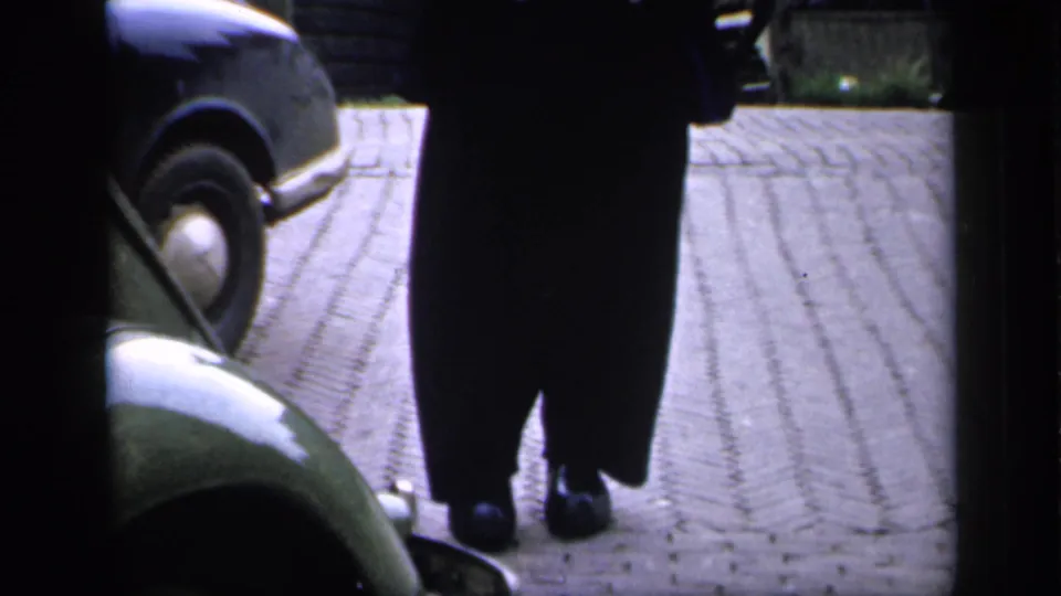 The lower half of a man in a long dark coat appears on an Irish cobblestone street with a rounded car at the frame edge.
