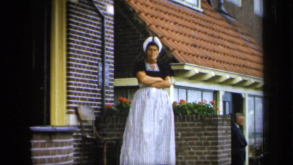 In 1949 Ireland, a woman in a white bonnet and long dress stands with arms crossed outside a brick and timber building with flowers nearby.