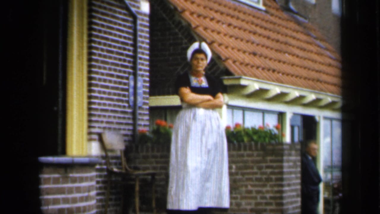 In Ireland 1949 footage, a woman in traditional-looking dress stands with arms crossed outside a brick house with flower boxes.