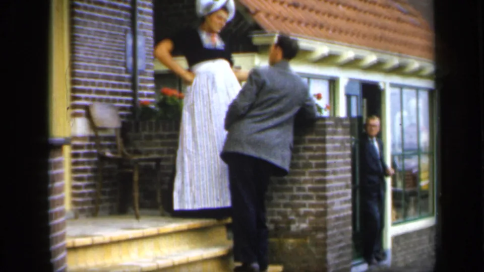 In Ireland 1949 footage, a woman in traditional-looking dress stands above a suited man near a brick house entrance.
