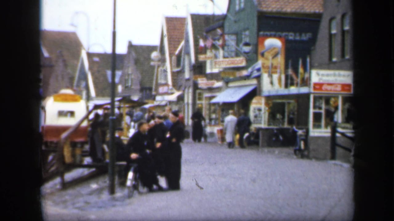 Cobblestone street with pedestrians, gabled shops, signs, and men gathered near a small vehicle or cart.