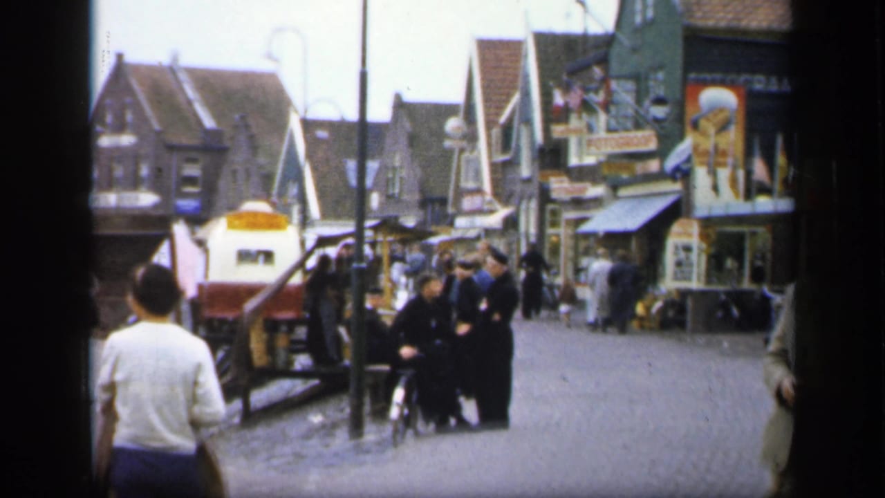 A man in a light shirt walks away from the camera as pedestrians gather near shops and street signs.