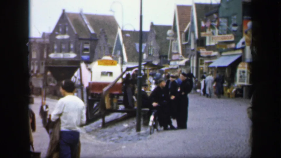 1949 Ireland home-movie frame showing a man from behind, shop-lined cobblestones, visible signs, and people conversing in the street.