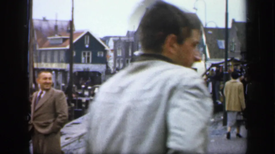 A man in a light jacket crosses close to the 8mm camera on a 1949 Irish street, with pedestrians and gabled shopfronts behind.