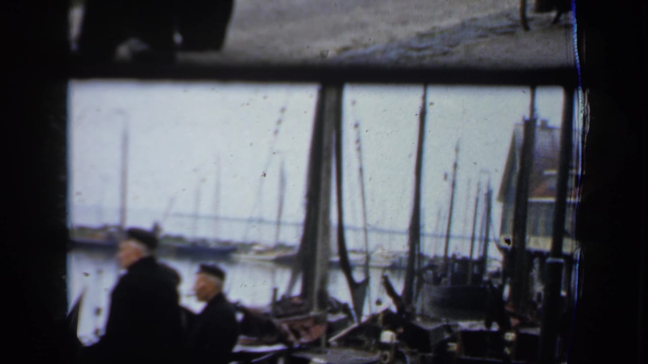 Two men in dark coats stand near boat rigging with sailboat masts across the water.