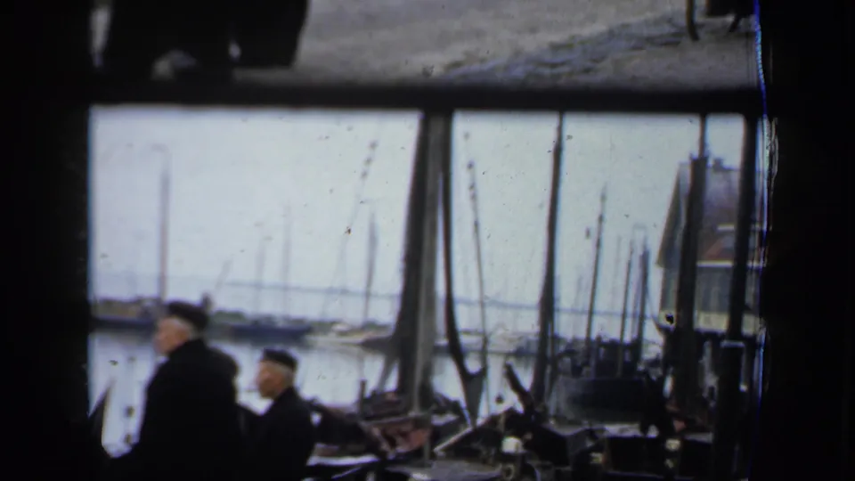 In 1949 Ireland 8mm footage, two men in dark coats and caps stand near a harbor with sailboat masts beyond them.