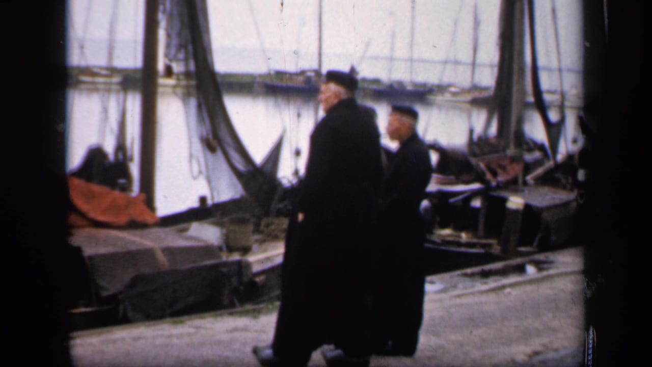 Two men in dark robes walk along a harbor dock beside boats and rigging.