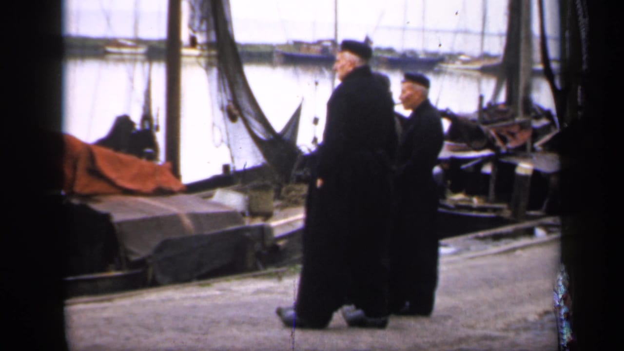 Two robed men continue walking on a dock with boats and masts behind them.