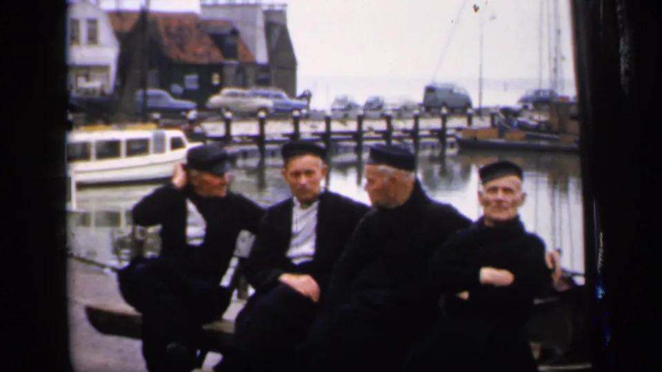 Four men in dark robes and caps sit together at an Irish harbor, with boats, parked cars, and quay buildings behind them.