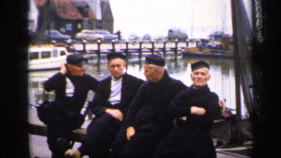 Four clerically dressed men sit at a quiet Irish harbor in 1949, framed by boats, water, parked cars, and dockside rigging.