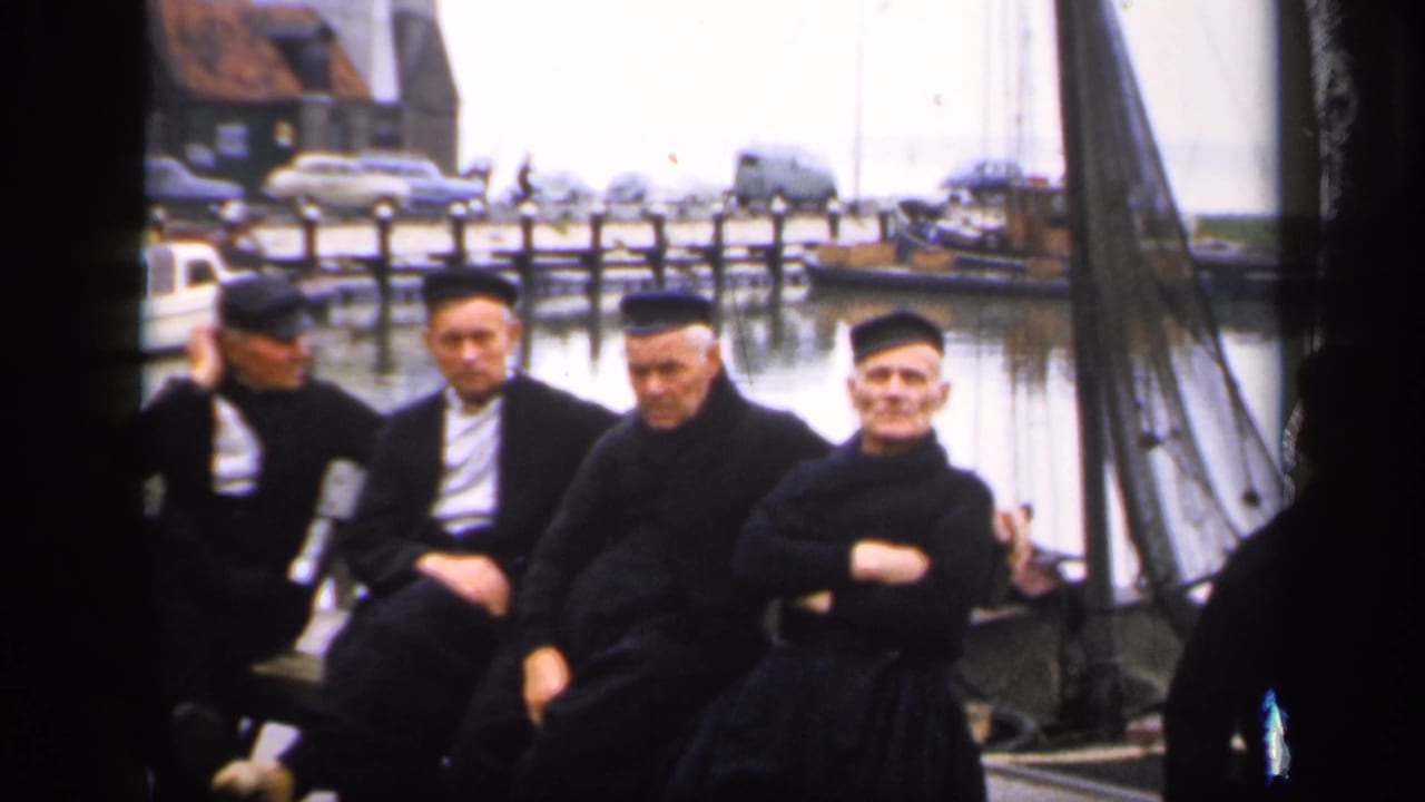 Five men in dark clothing sit on a dock beside harbor boats.