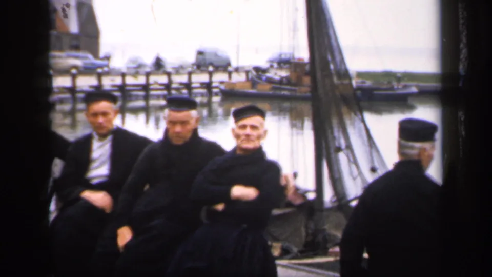 1949 Ireland dockside frame with older men in dark clothing seated near a sailboat while another capped man faces the harbor.