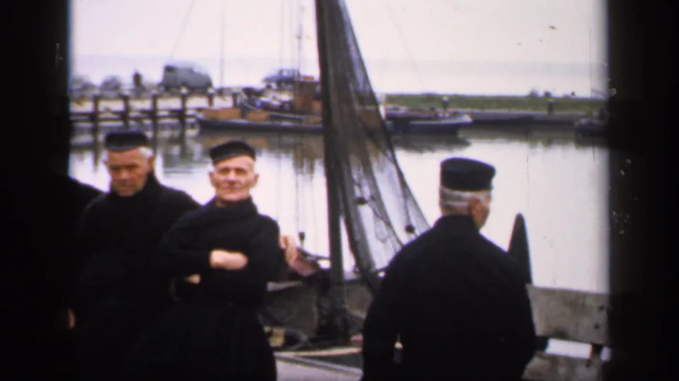 1949 Ireland harbor frame showing two seated older men and a capped man seen from behind beside a docked sailboat.