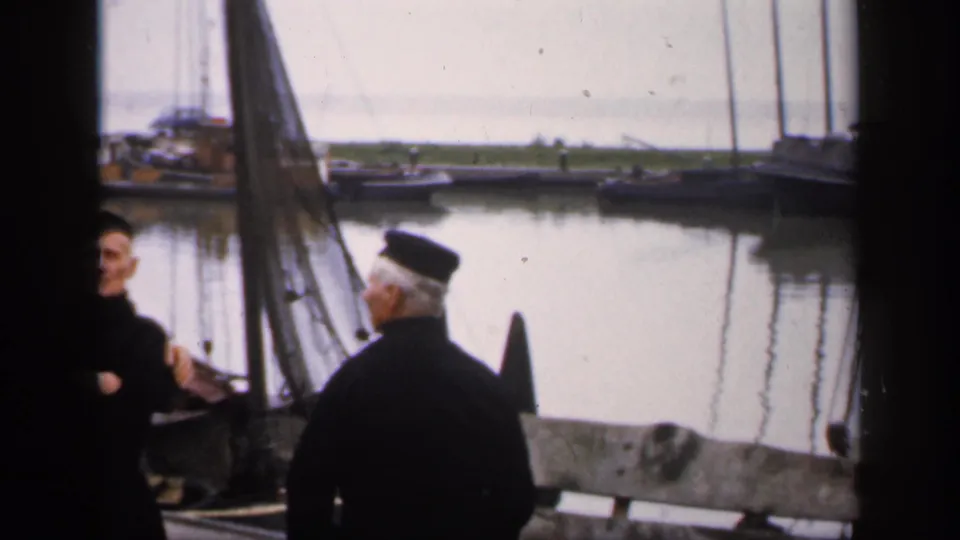 1949 Ireland home-movie frame with a man seen from behind on the dock, sail rigging nearby, and small boats across calm water.