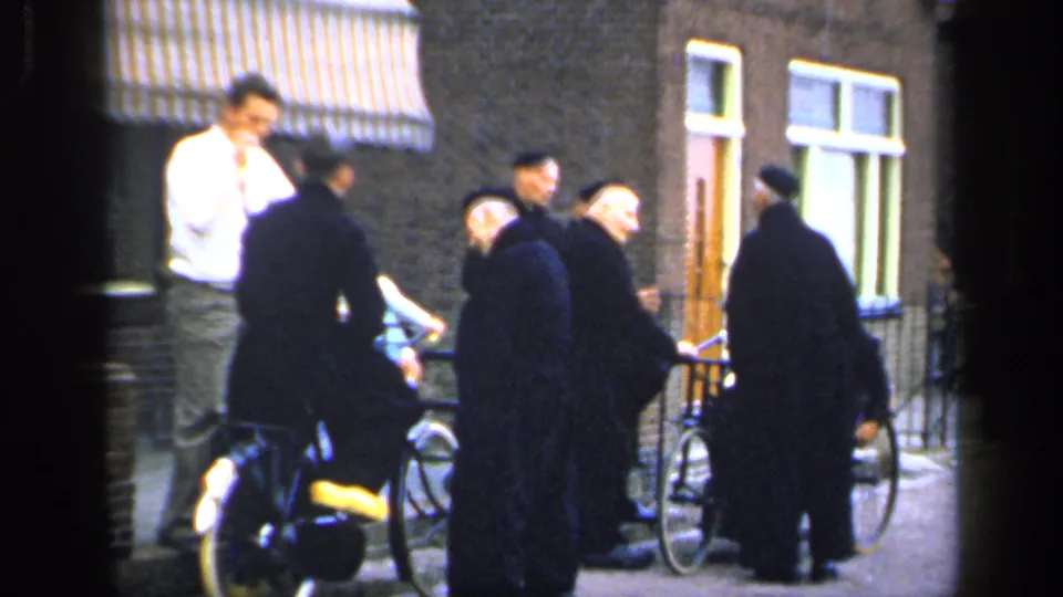 In 1949 Ireland, men in dark coats or robes gather with bicycles on a street beside brick facades, windows, and a striped awning.