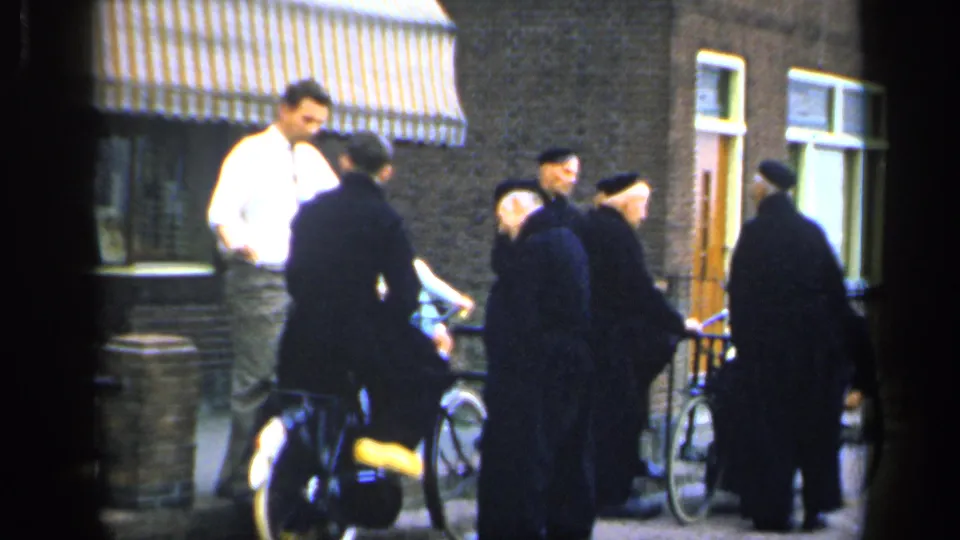 The 1949 Ireland home-movie frame shows a young man in a white shirt and tie near older men in dark clothing with bicycles outside brick buildings.