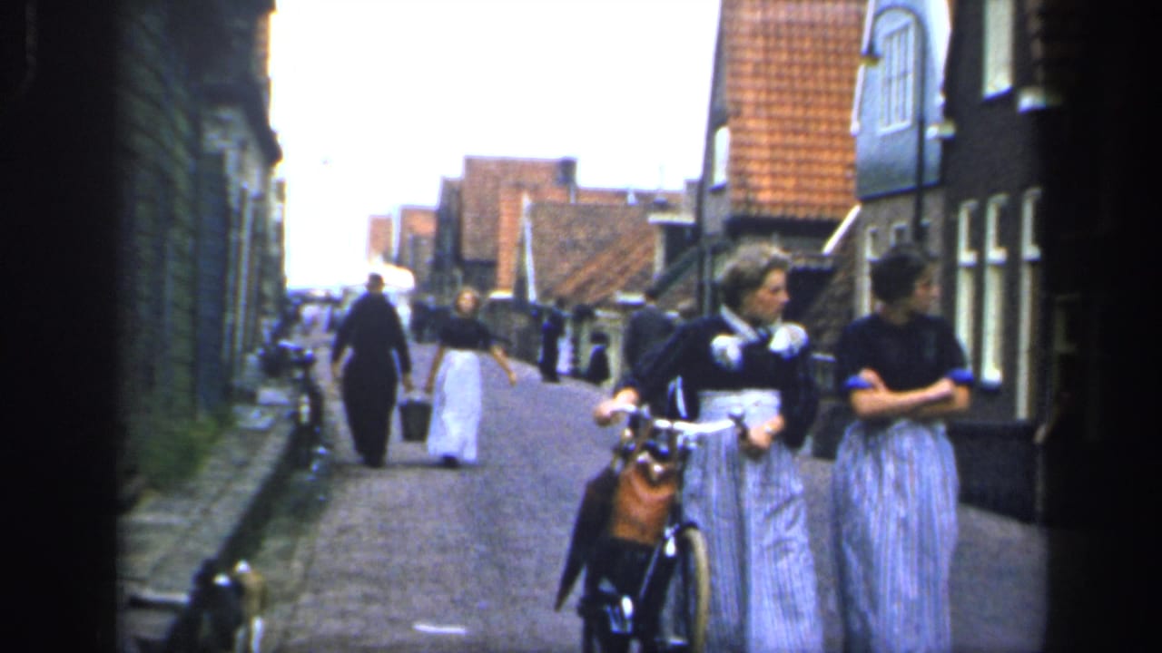 In Ireland 1949 home-movie footage, women in long striped skirts walk along a narrow cobbled street while one holds a bicycle.