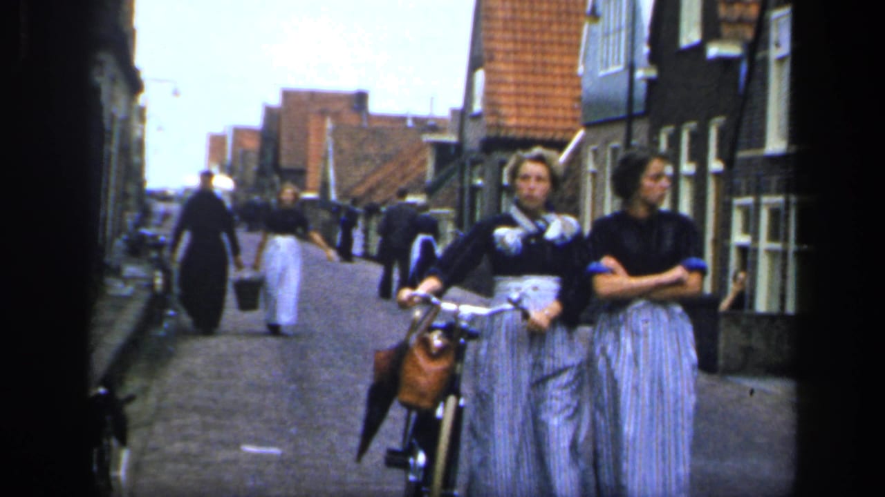 Two women in long skirts pause beside a bicycle on an Irish town street, with other people carrying buckets in the background.