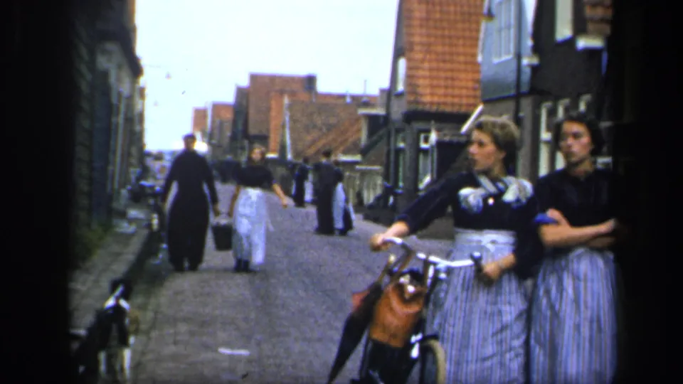 A handheld 8mm frame shows women moving along a cobbled Irish street, including one near a bicycle at the foreground edge.