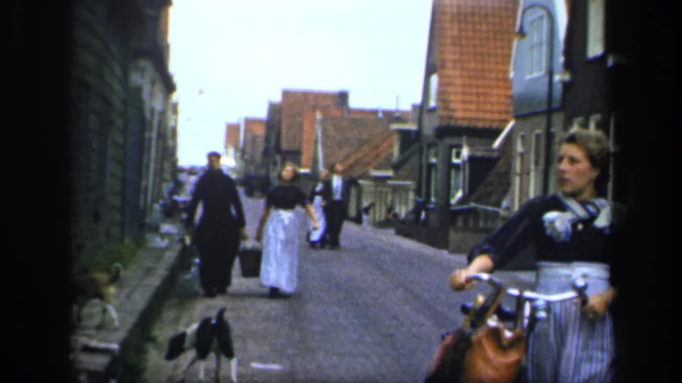 A woman holding a bicycle is framed at the edge of a 1949 Ireland street scene, with pedestrians and a dog farther along the road.