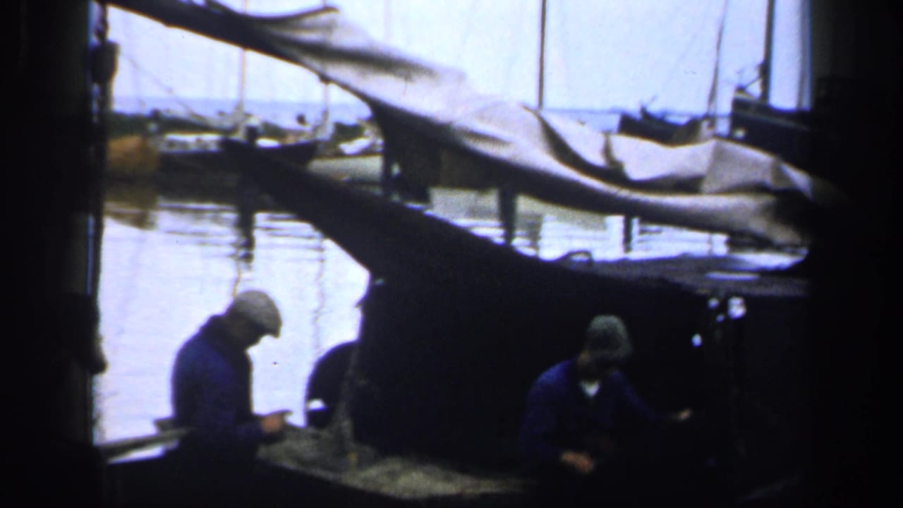 1940s Ireland harbor view with fishermen in work clothes handling gear beside a canvas-covered boat and calm water.