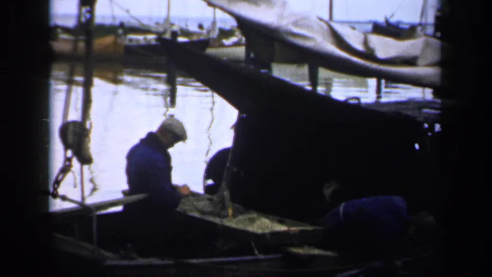 1940s Ireland harbor frame showing a fisherman standing over gear while another bends near nets beside a dark boat.