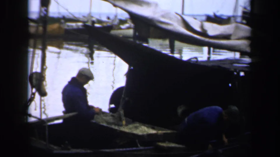 1940s Ireland home-movie frame of harbor workers handling nets or gear near a moored boat with other vessels beyond.