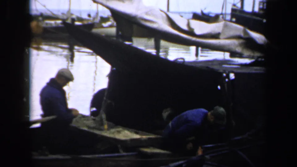 1949 Ireland home-movie frame showing two men working aboard a small harbor boat, with covered boats and water behind them.