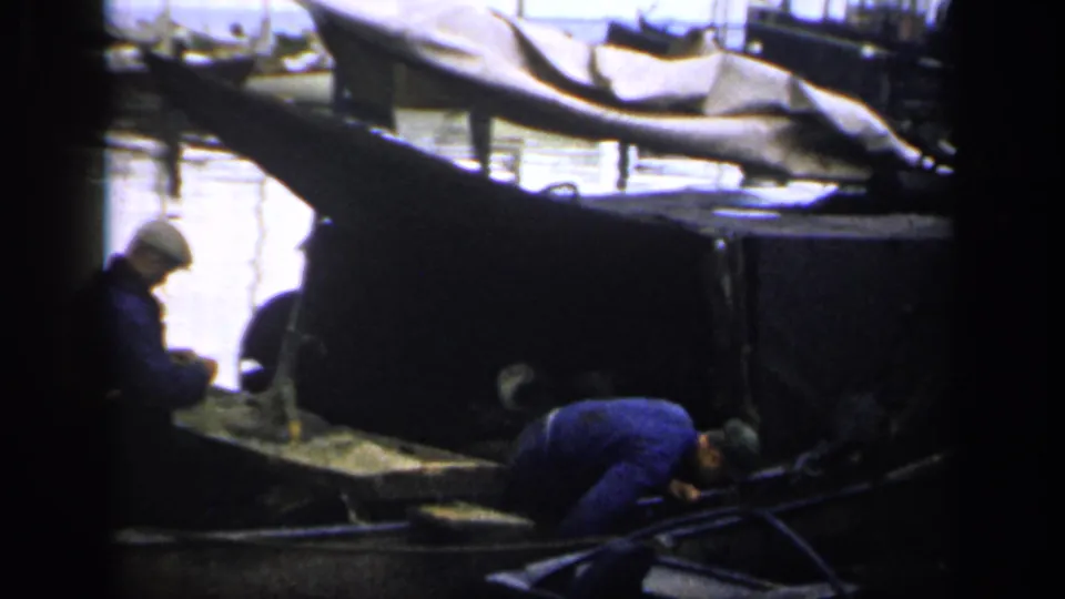 The home-movie frame shows two men in blue clothing maintaining or handling something in a small boat at an Irish harbor.