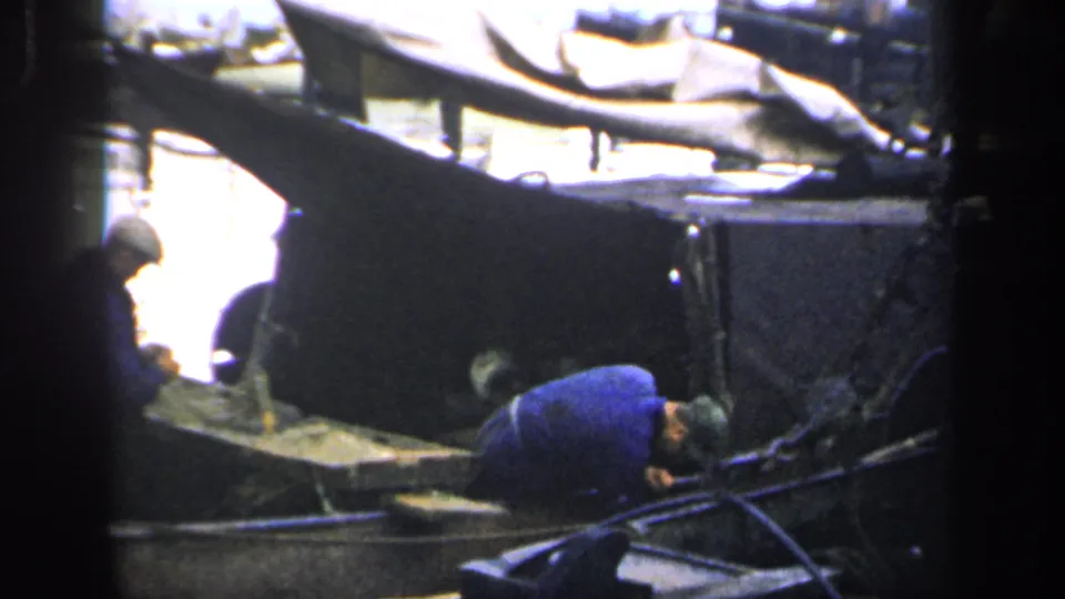 A 1949 Ireland harbor frame shows one worker bent low inside a small boat and another seated beside him, with moored boats behind.