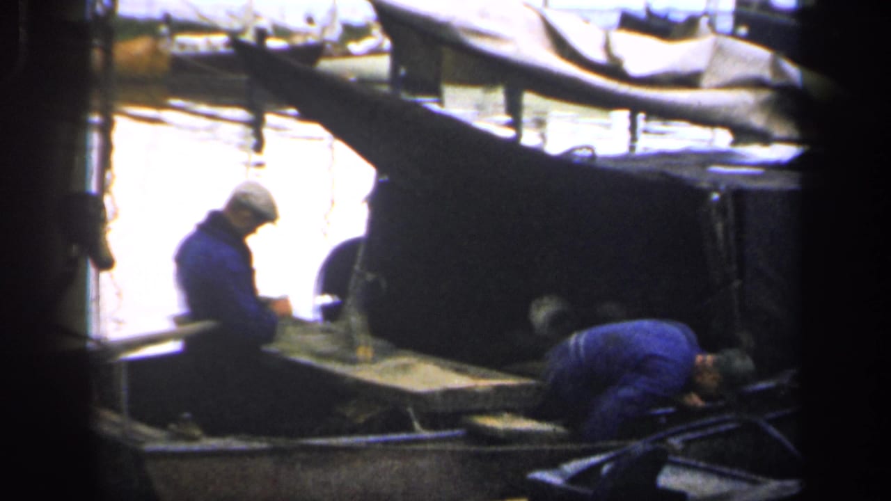 Two men work inside a small boat beside calm harbor water.