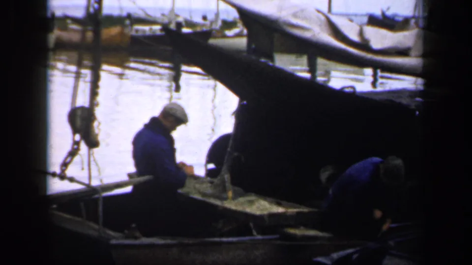 A 1949 Ireland waterfront frame shows one man standing and another bent over in a small fishing boat beside moored, covered boats.