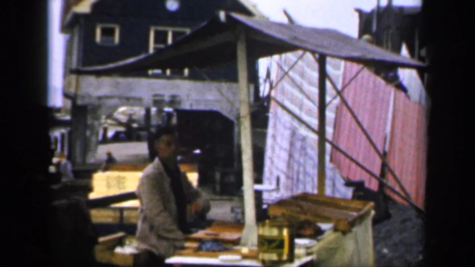 A 1949 Ireland village waterfront view shows a man seated under a tarp at a makeshift outdoor table, with laundry and buildings behind him.