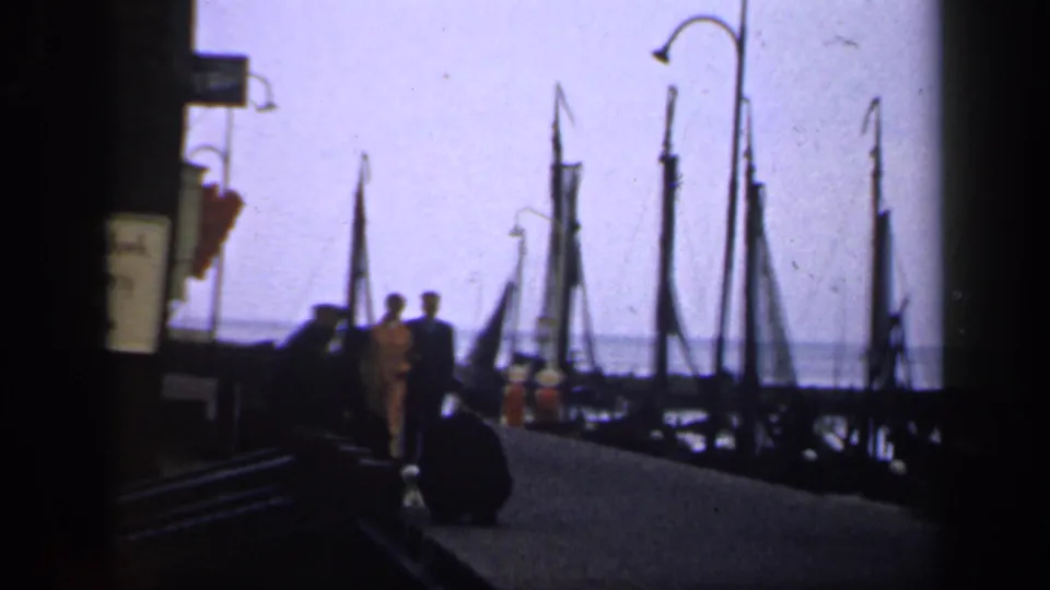 In Ireland around 1949, silhouetted adults walk on a dock past sailboats, curved lamps, and calm water.