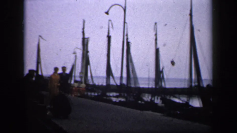 The couple appears farther along the Irish dock, framed by moored sailboats, upright masts, and a pale sky.